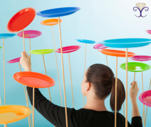 A woman stands in the middle of a number of colored plates spinning on sticks. She is trying to control them to keep them all spinning. 